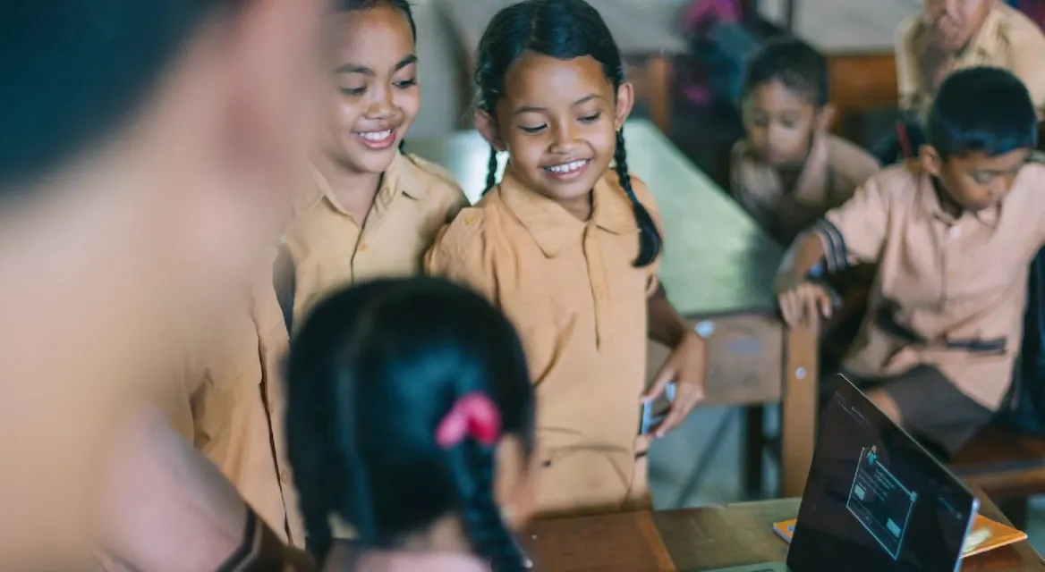 Children working around a laptop in a busy classroom.