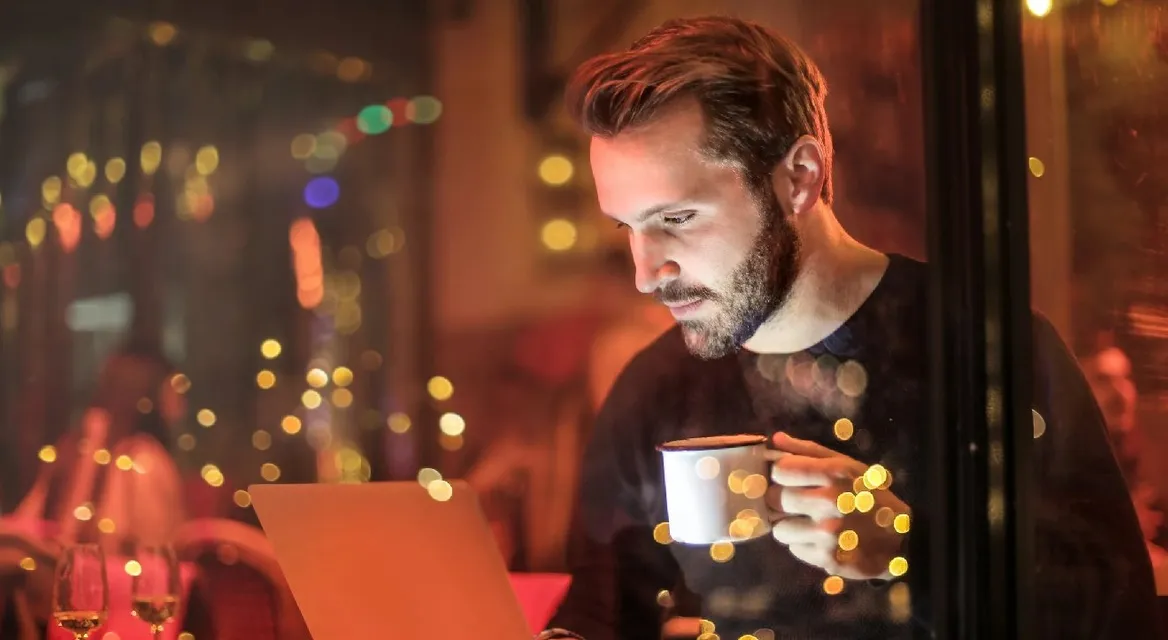 A man in front of a laptop with a coffee in a dimly lit cafe.