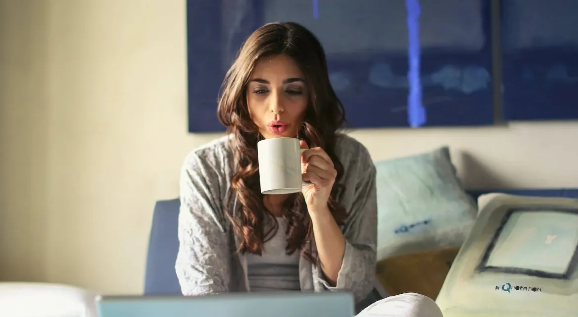 A woman drinking a cup of coffee in bed in front of her laptop.