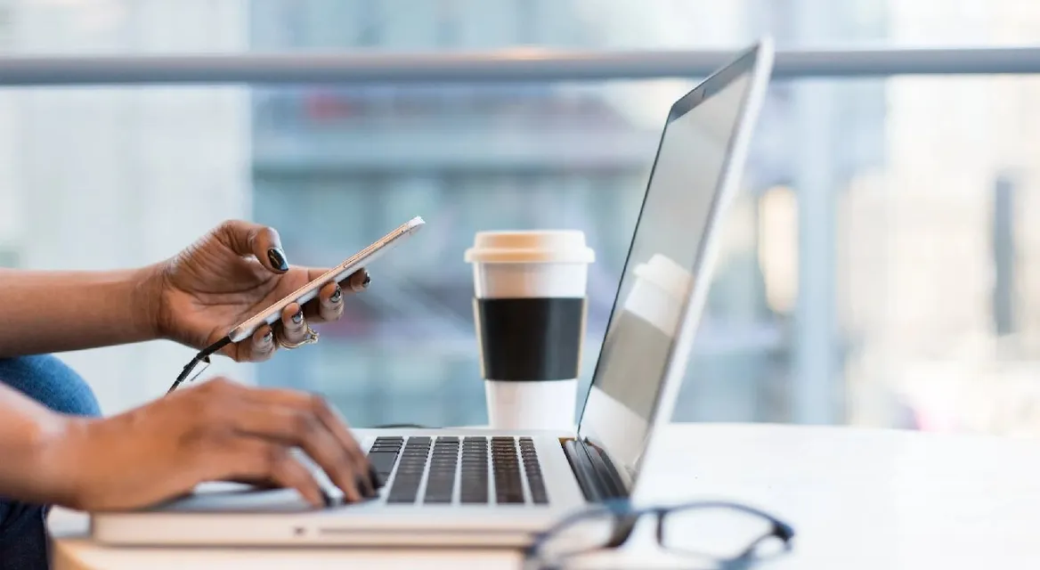 A side-on profile of someone working on a laptop with a phone in their hand, and a coffee next to them.