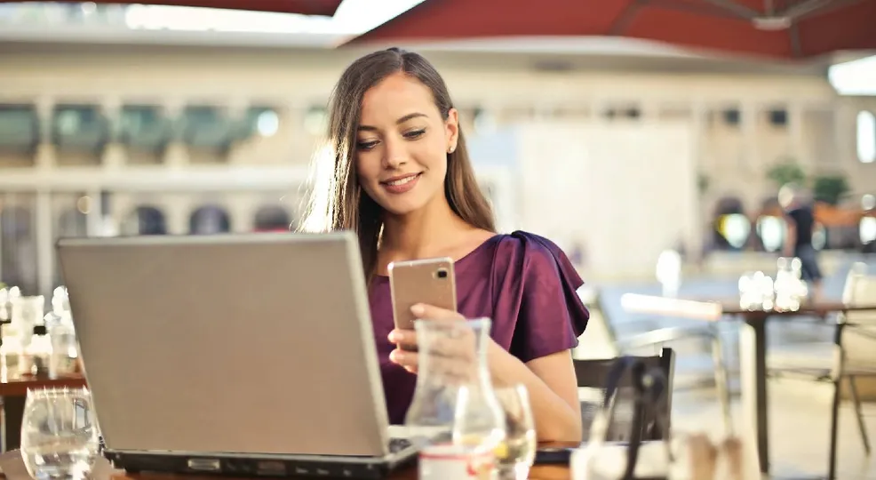 A woman on her phone in front of a laptop.