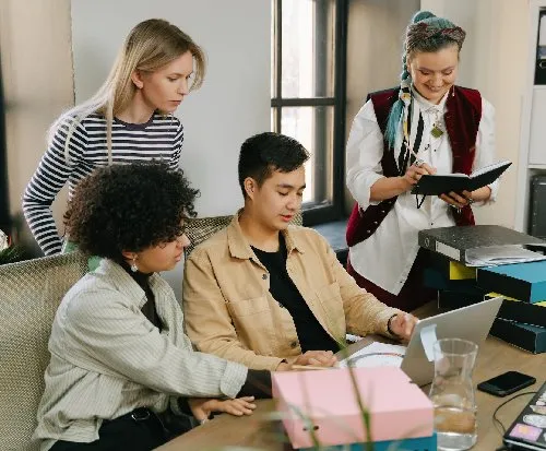A group of four people working in front of one laptop in an office environment.